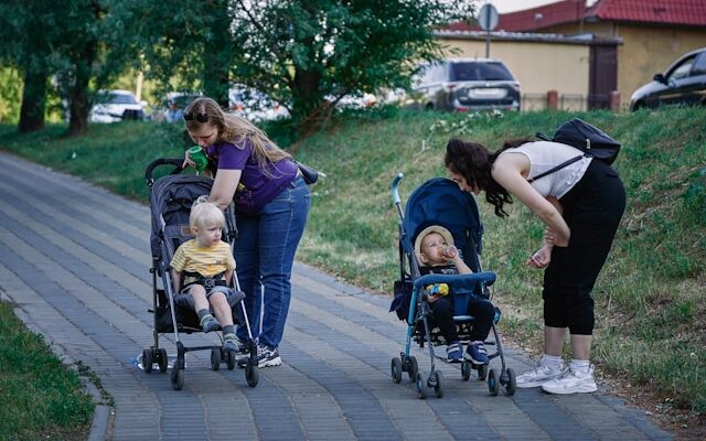 Two women stand on a sidewalk with toddlers seated in strollers.