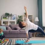 A mom doing somatic exercise in a side angle pose on a blue mat in the living room while her young child plays with colorful blocks on a striped rug