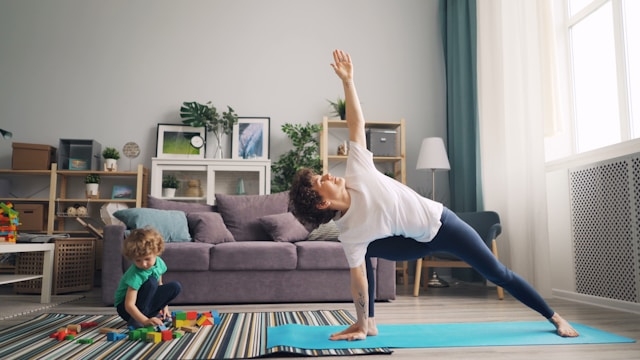 A mom doing somatic exercise in a side angle pose on a blue mat in the living room while her young child plays with colorful blocks on a striped rug