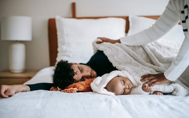 A father and his baby sleeping together on a bed, covered with a soft white blanket, while another person gently tucks them in.