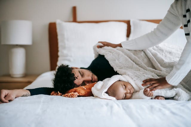A father and his baby sleeping together on a bed, covered with a soft white blanket, while another person gently tucks them in.