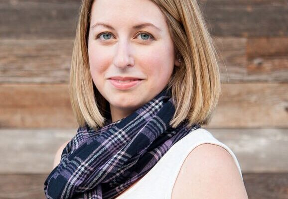 Kellie, a stylish mom and founder of The Yuppie Files lifestyle blog, wearing a plaid scarf and white top, smiling against a wooden background.