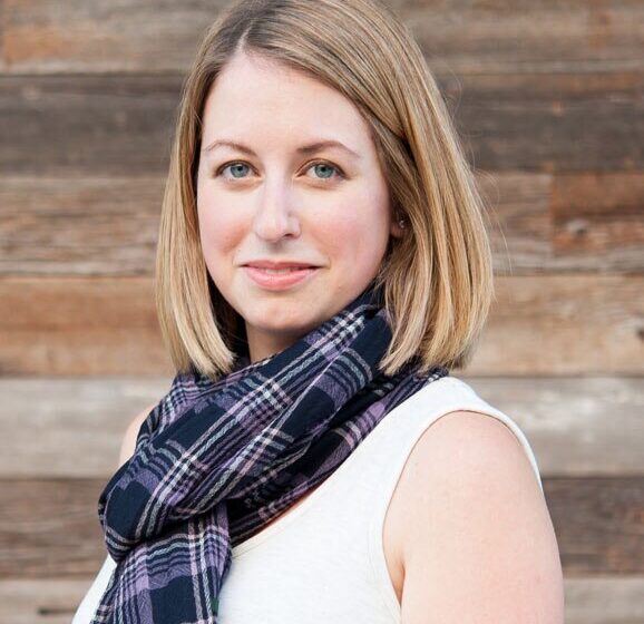 Kellie, a stylish mom and founder of The Yuppie Files lifestyle blog, wearing a plaid scarf and white top, smiling against a wooden background.