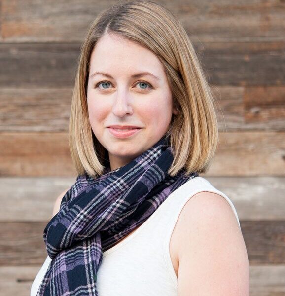 Kellie, a stylish mom and founder of The Yuppie Files lifestyle blog, wearing a plaid scarf and white top, smiling against a wooden background.
