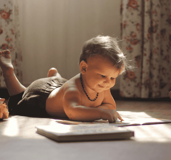 Smiling toddler lying on the floor and pointing at a book, symbolizing early learning and positive behavior support.