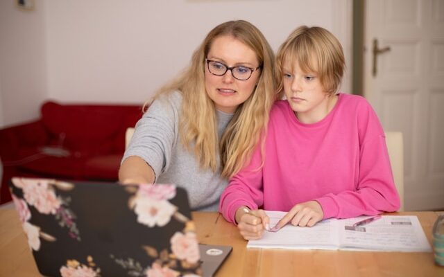A woman wearing glasses teaching a child with schoolwork, pointing at a laptop while the child writes in a notebook.