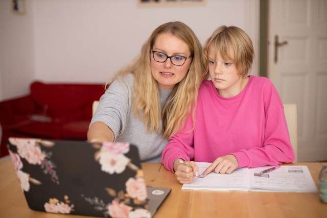 A woman wearing glasses teaching a child with schoolwork, pointing at a laptop while the child writes in a notebook.