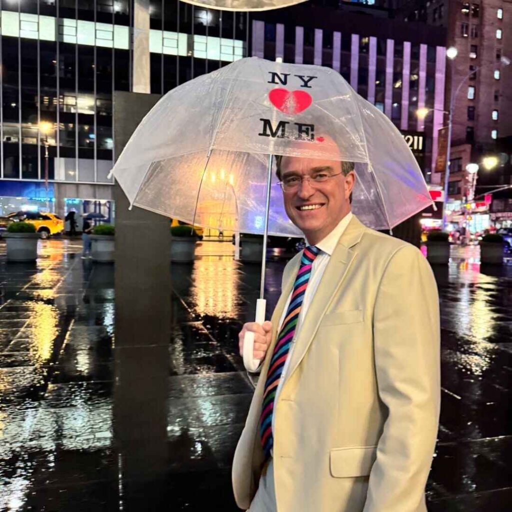 Charles Hurt with an umbrella in New York Charles Hurt smiling and holding a "NY ♥ ME" umbrella in Times Square at night with rain reflecting the city lights.