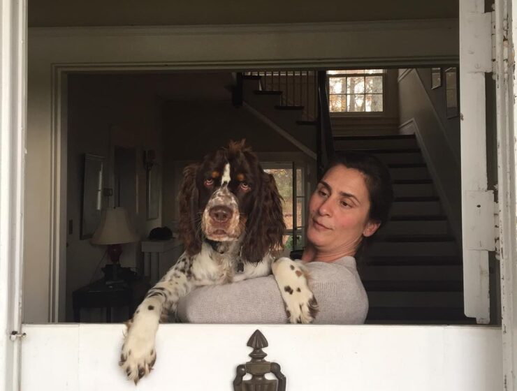 Stephanie Hurt, wife of Charles Hurt, standing by a half-open door and holding a brown-and-white dog, with a staircase visible in the background.