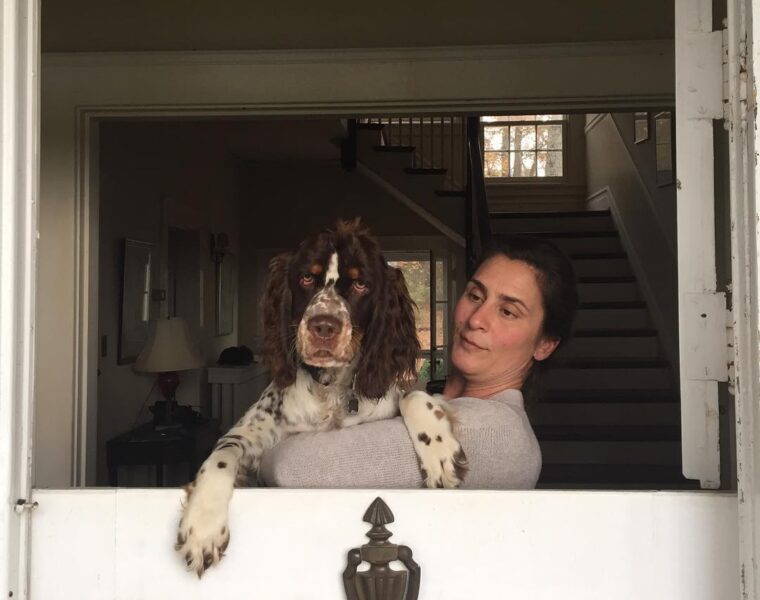 Stephanie Hurt, wife of Charles Hurt, standing by a half-open door and holding a brown-and-white dog, with a staircase visible in the background.