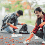 A young man and woman wearing orange gloves crouching on a paved path to pick up plastic bottles and litter during a community cleanup event.