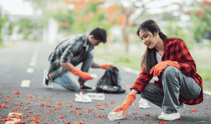 A young man and woman wearing orange gloves crouching on a paved path to pick up plastic bottles and litter during a community cleanup event.