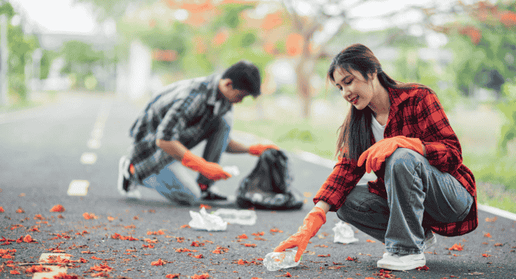 A young man and woman wearing orange gloves crouching on a paved path to pick up plastic bottles and litter during a community cleanup event.