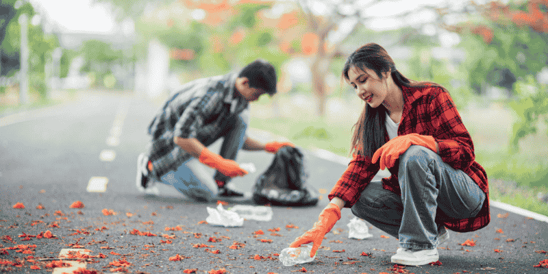 A young man and woman wearing orange gloves crouching on a paved path to pick up plastic bottles and litter during a community cleanup event.