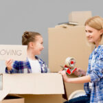 Mother and child packing boxes together, with the child holding a “moving” sign, illustrating a positive and calm approach to discussing relocation with kids.
